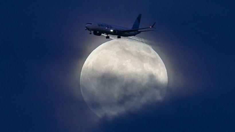 A large, glowing white moon rises above blue clouds, a plane flying alongside it near San Francisco Bay